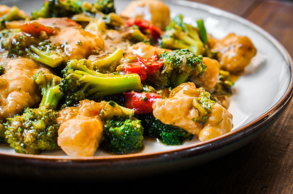 A plate of stir-fried chicken with vibrant broccoli and red bell peppers is served on a wooden table, showcasing a glossy sauce that enhances the colorful vegetables and tender chicken pieces.