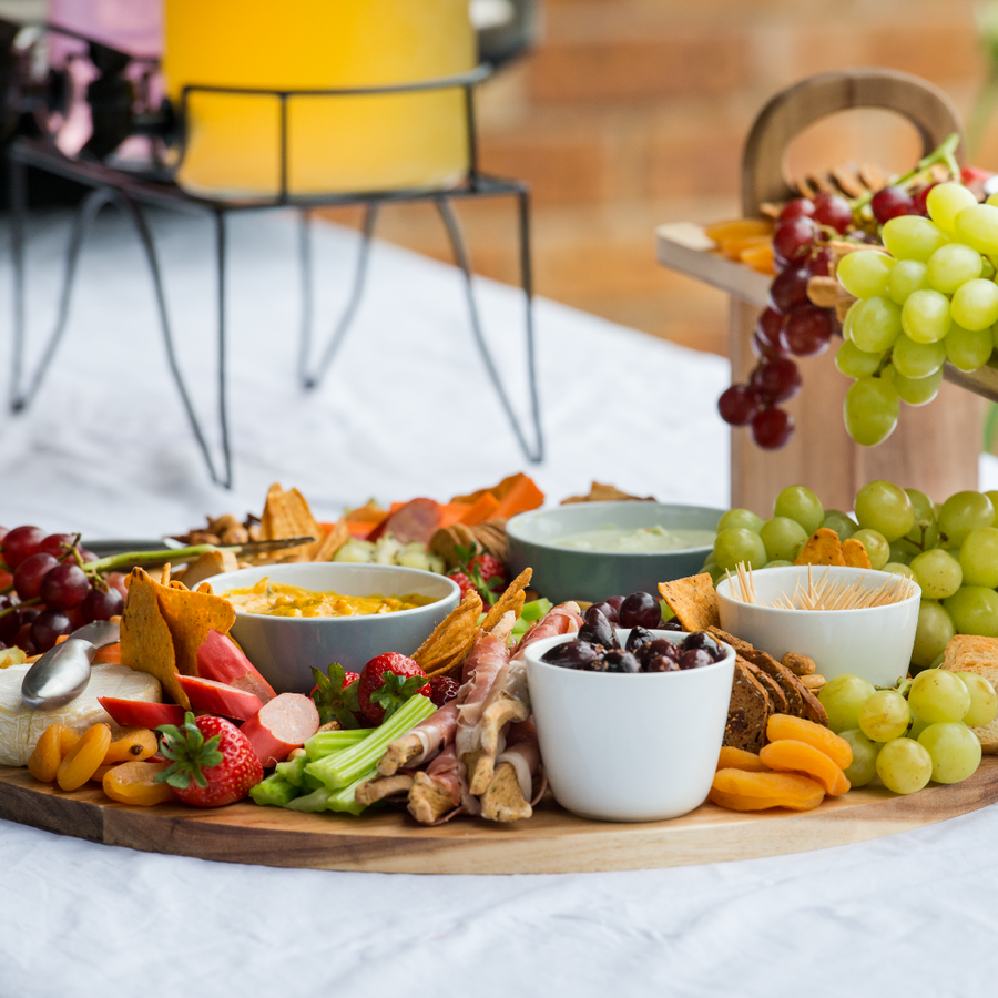 A wooden platter displays a variety of colorful summer snacks, including fresh fruits, vegetables, dips, and crackers, arranged attractively on a white tablecloth with a drink dispenser in the background.