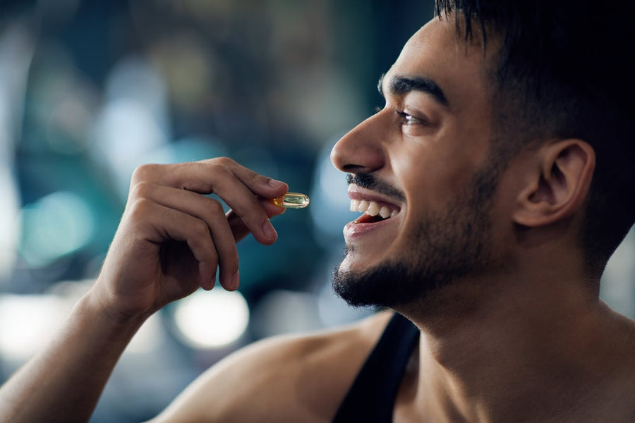 A smiling man holds a nutritional supplement capsule in his hand, preparing to take it, with a blurred gym setting in the background, emphasizing the importance of supplements after weight loss surgery.