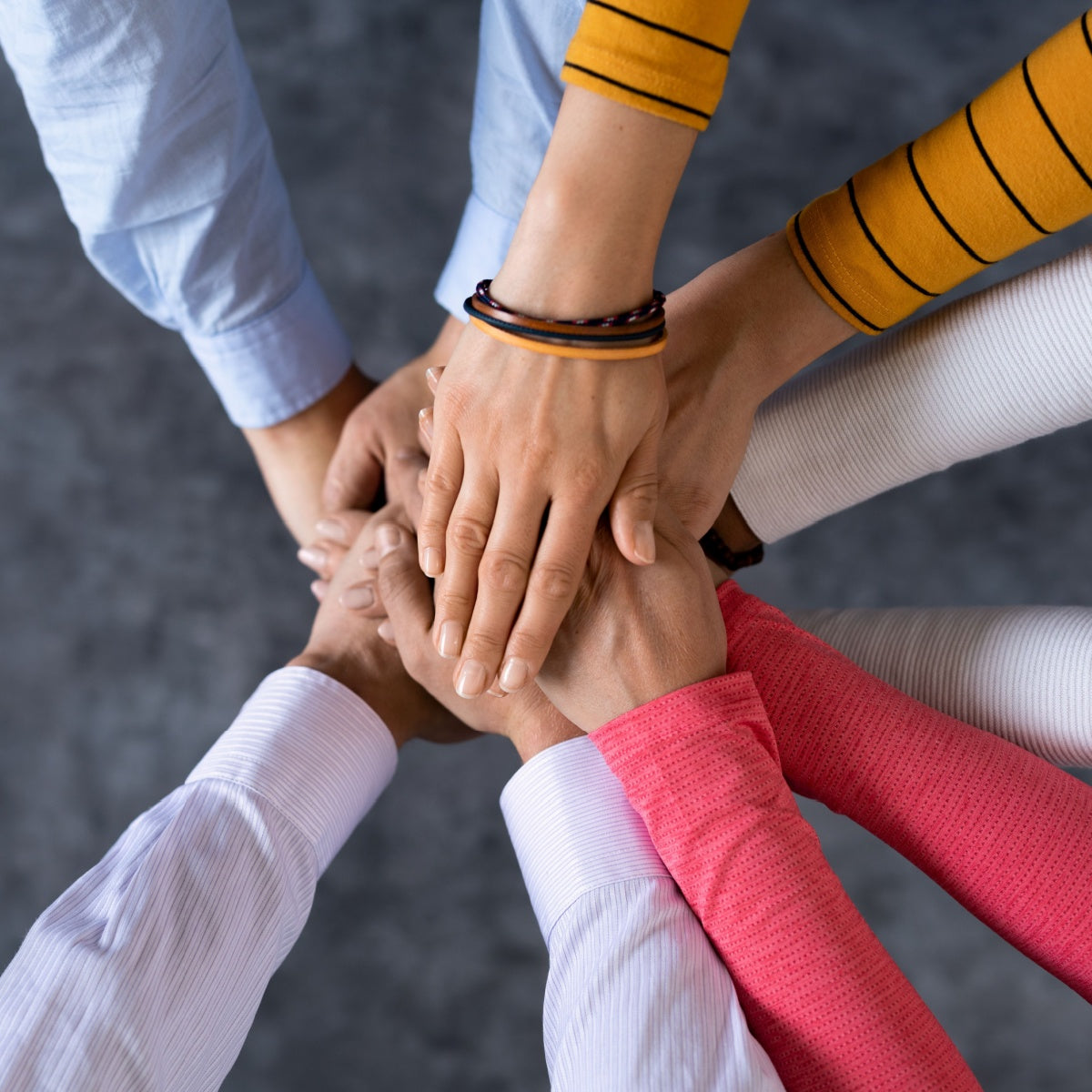 Multiple hands of diverse individuals are stacked together in a supportive gesture, symbolizing unity and collaboration, set against a blurred gray background.