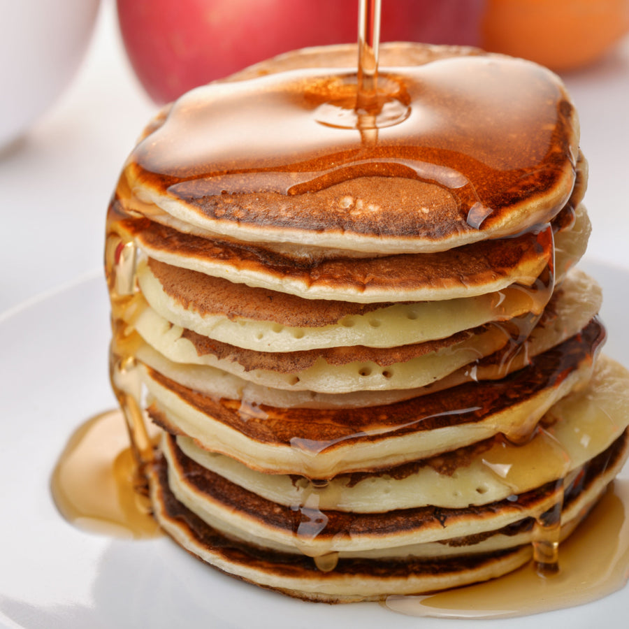 A stack of golden-brown pancakes is drizzled with syrup, sitting on a white plate, with apples and an orange blurred in the background.