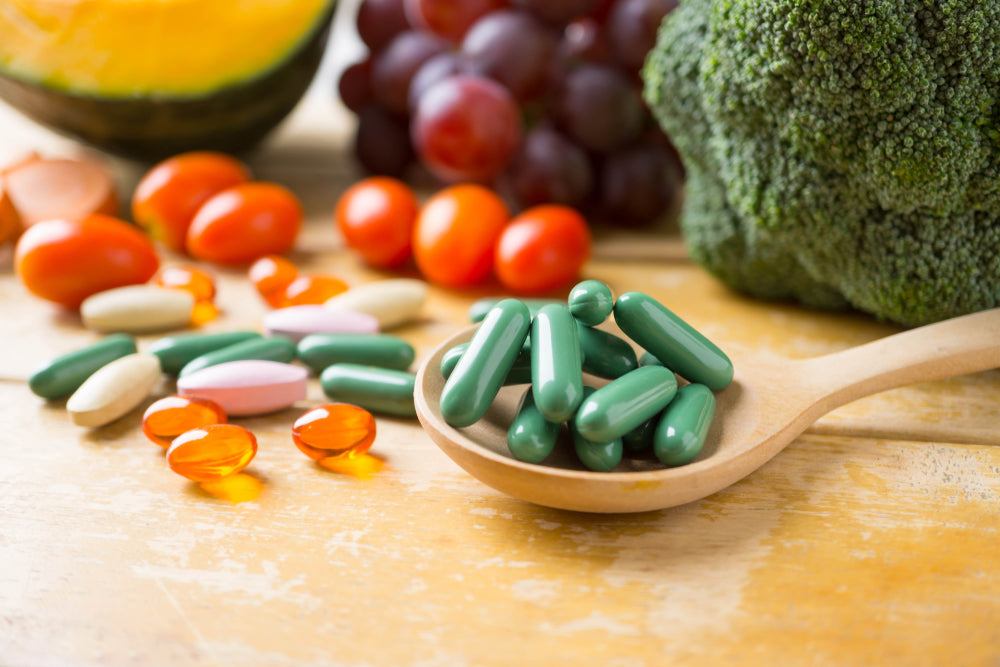 A wooden spoon holds green capsules among various vitamins and supplements scattered on a table, with colorful fruits and vegetables like broccoli and grapes in the background, emphasizing healthy nutrition post-bariatric surgery.