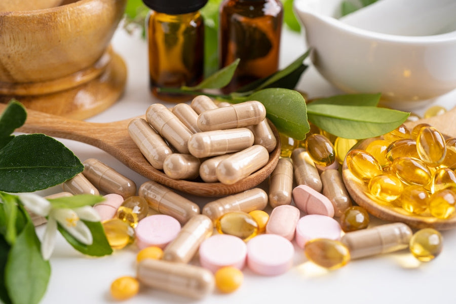 A wooden spoon holds beige capsules among various vitamins and supplements, with green leaves and small bottles in the background, suggesting a focus on health and nutrition.