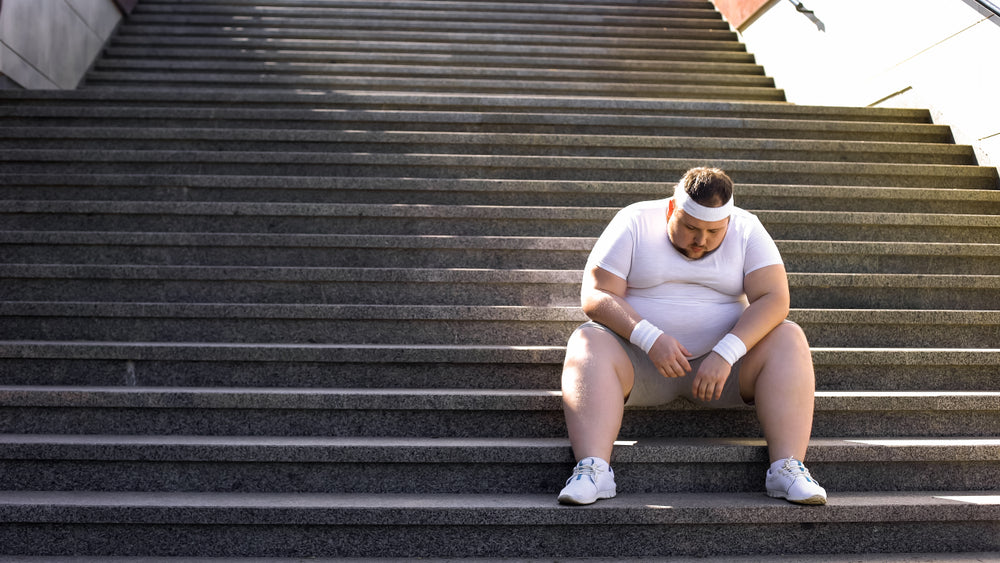 A man in a white workout outfit sits on outdoor steps, looking down with a tired expression, reflecting on his weight loss journey and health goals.