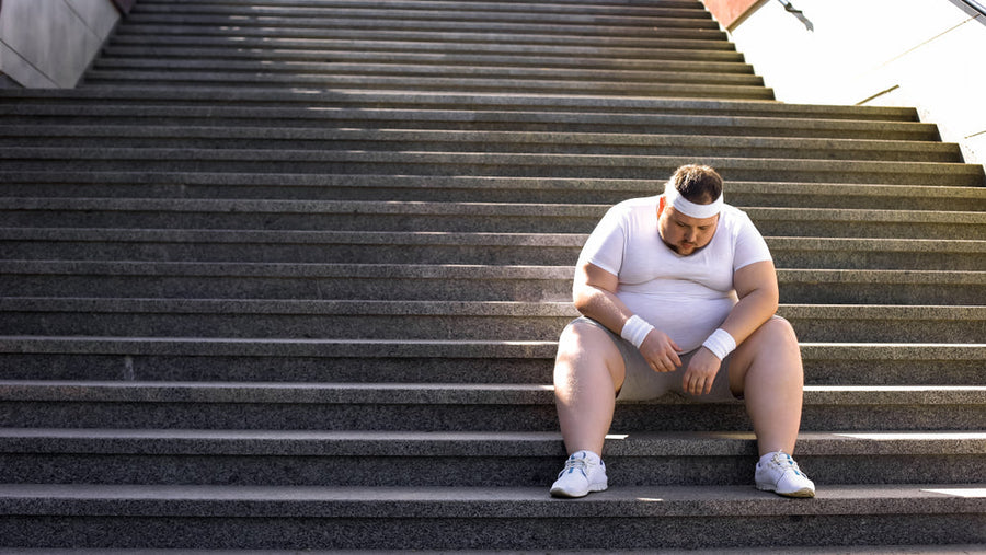 A man in a white workout outfit sits on outdoor steps, looking down with a tired expression, reflecting on his weight loss journey and health goals.