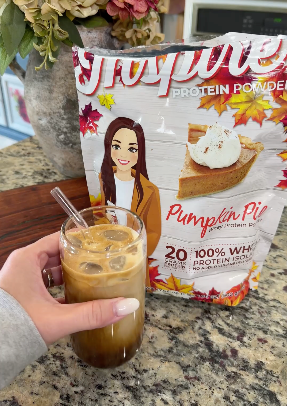 A hand holds a glass of iced coffee with a straw, next to a bag of pumpkin pie flavored protein powder decorated with autumn leaves on a kitchen countertop.