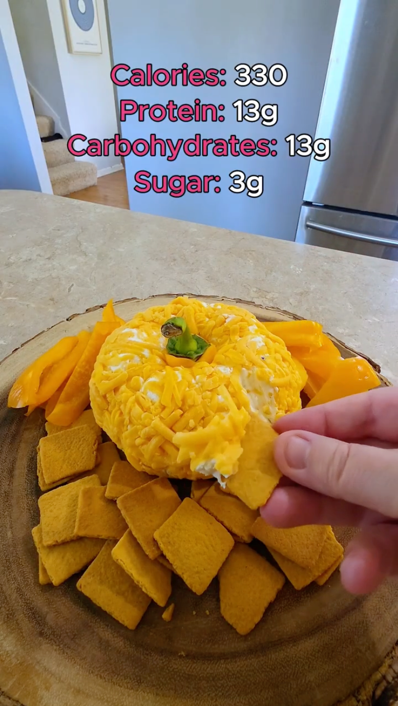 A hand reaches for a chip while a cheese ball shaped like a pumpkin sits on a wooden platter, surrounded by yellow bell pepper slices and crackers, with nutritional information displayed above.