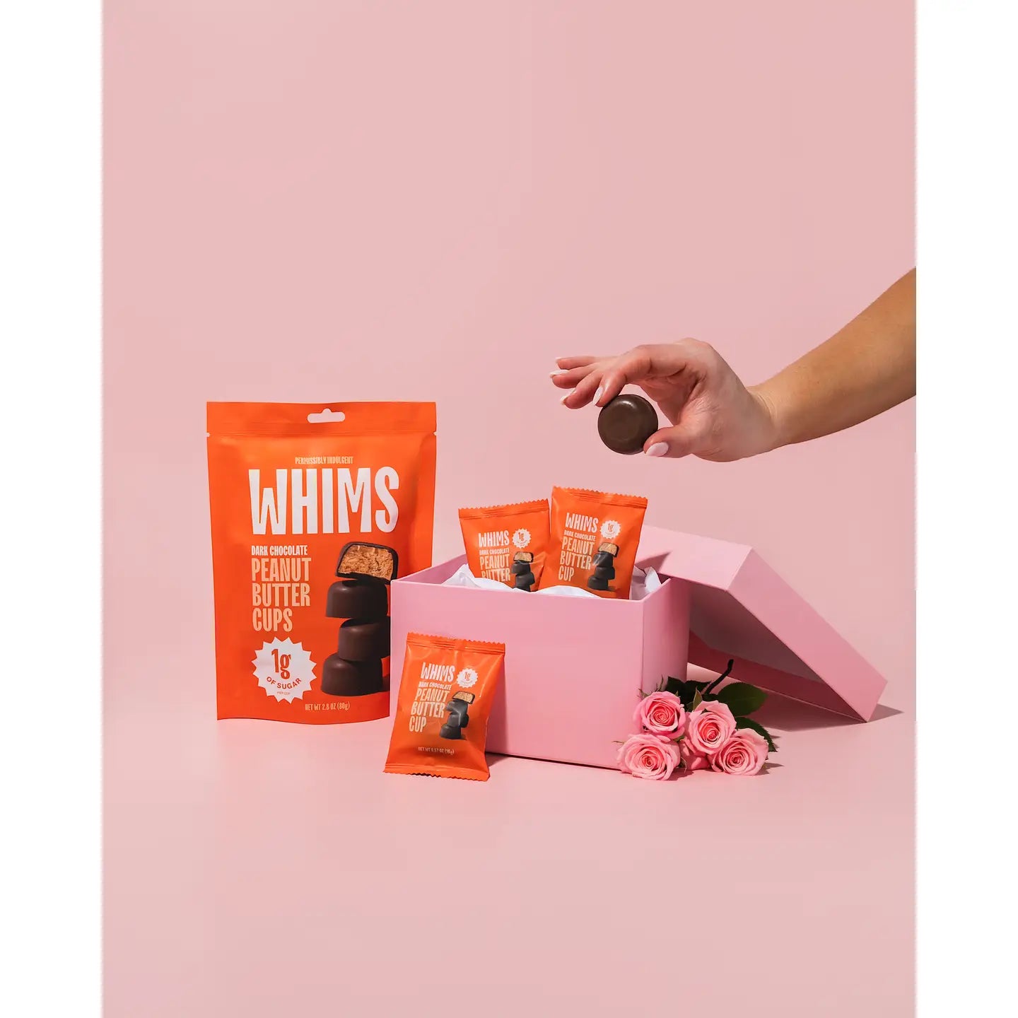 A hand reaches for a dark chocolate peanut butter cup from a pink box filled with individual packets of the same treat, set against a soft pink background with roses nearby.