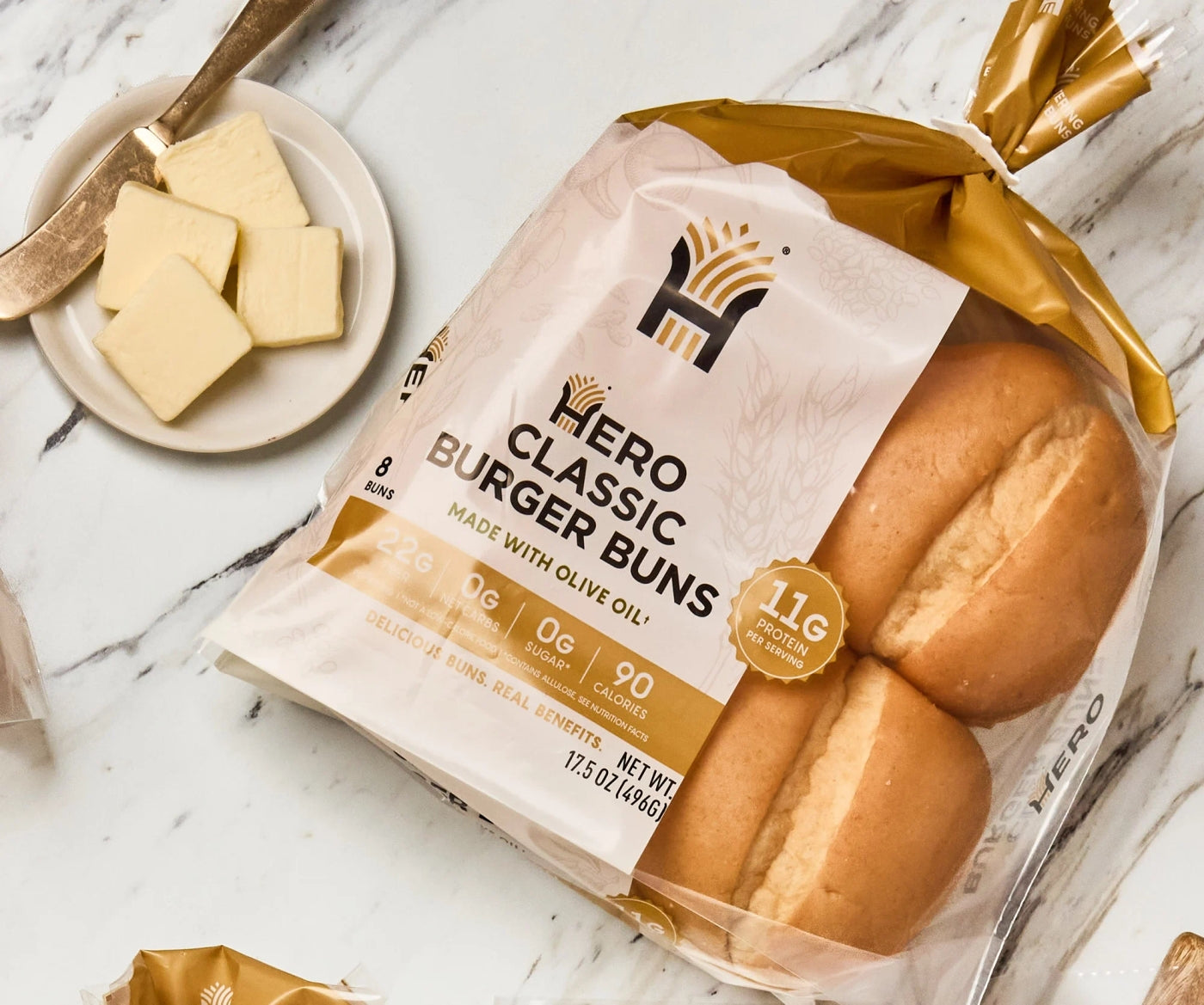 A package of Hero Classic Burger Buns sits on a marble surface next to a plate with butter slices, highlighting the product's nutritional information and branding.