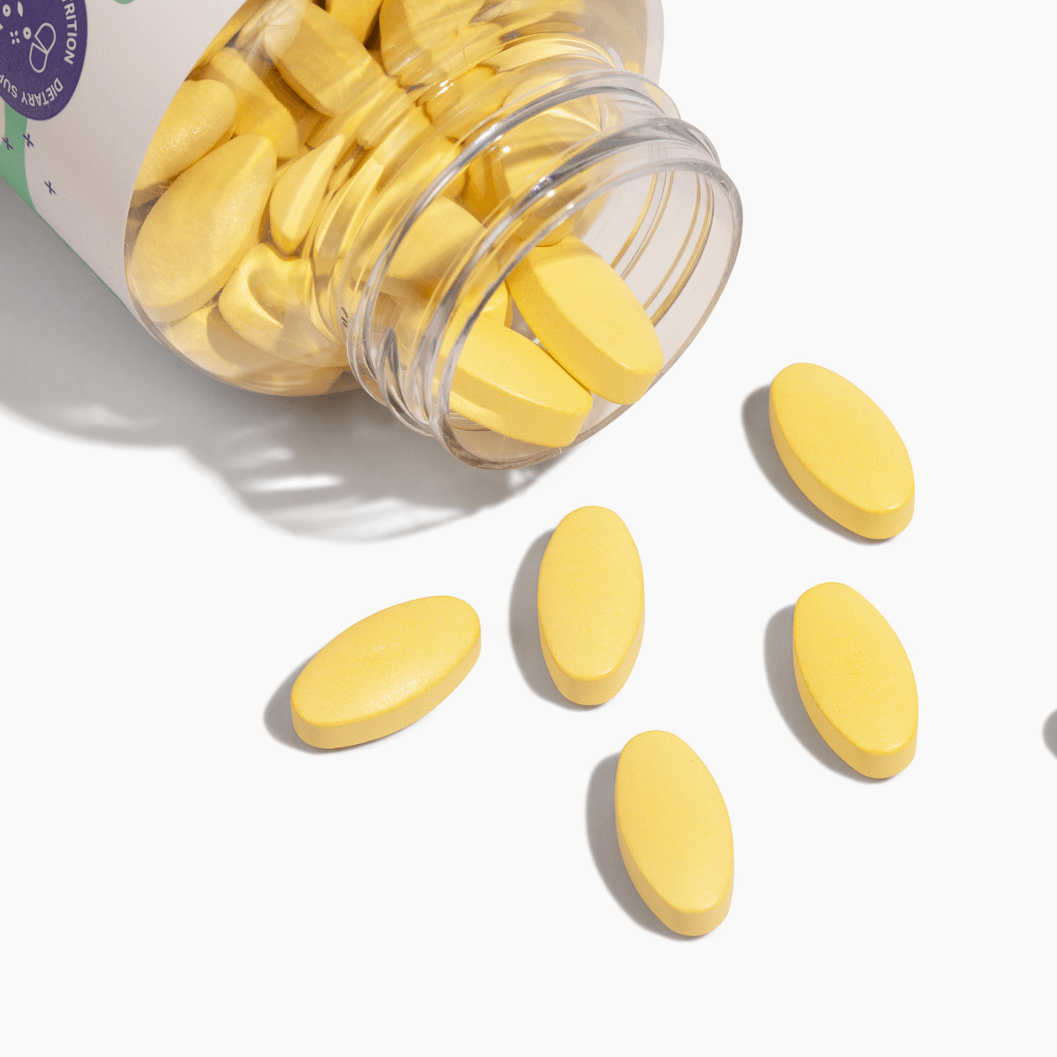 A clear bottle spills yellow oval vitamins onto a white surface, showcasing the product's vibrant color and texture.