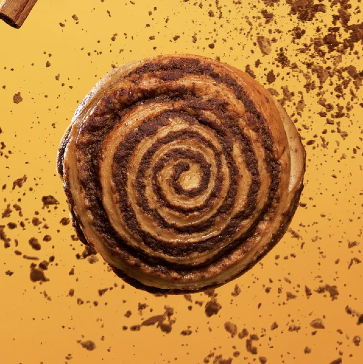 A freshly baked cinnamon roll with a golden-brown spiral sits on a vibrant orange background, surrounded by scattered cinnamon crumbs.