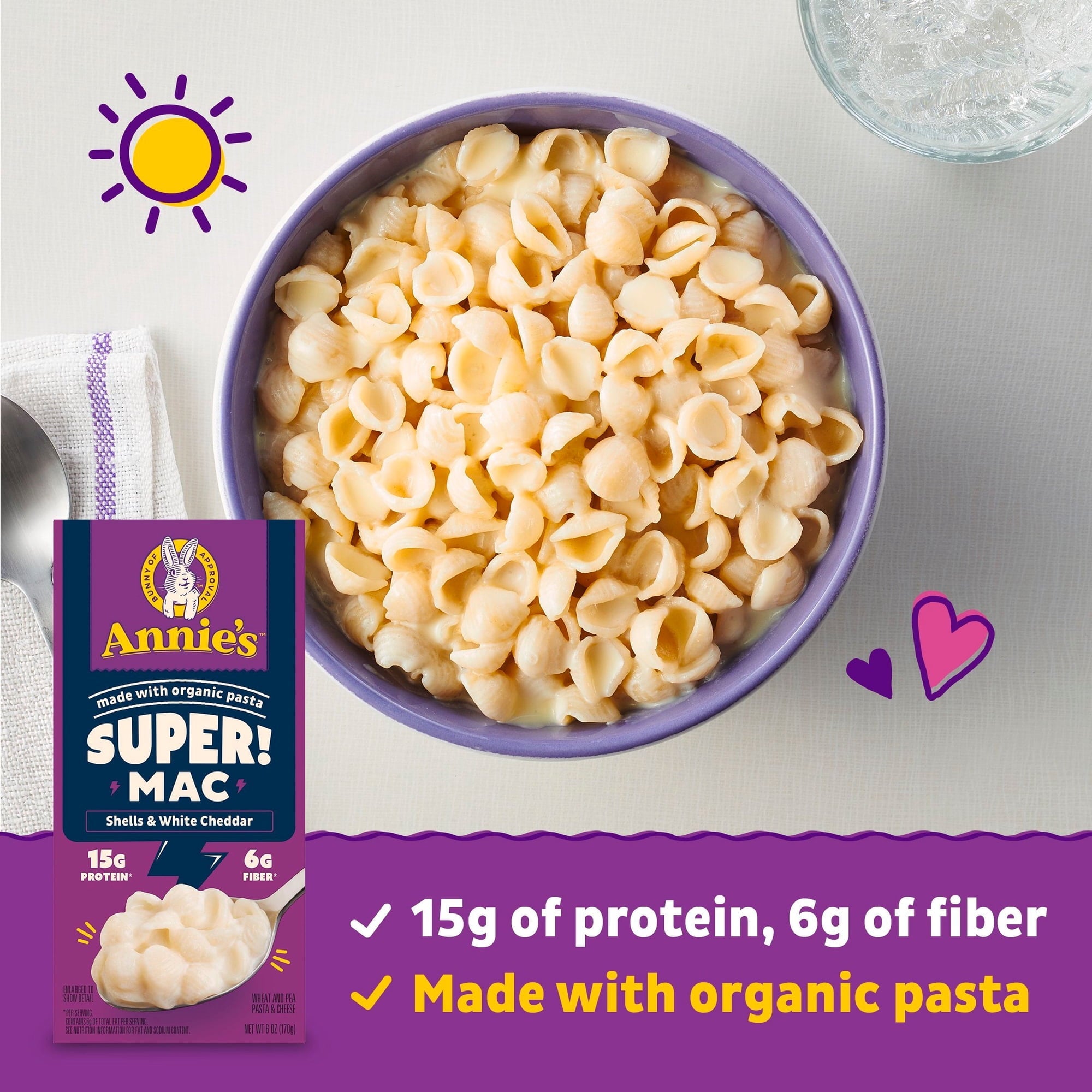 A purple bowl filled with creamy shells and white cheddar macaroni is placed on a light surface next to a spoon, with a box of Annie's Super Mac featuring nutritional highlights in the foreground.