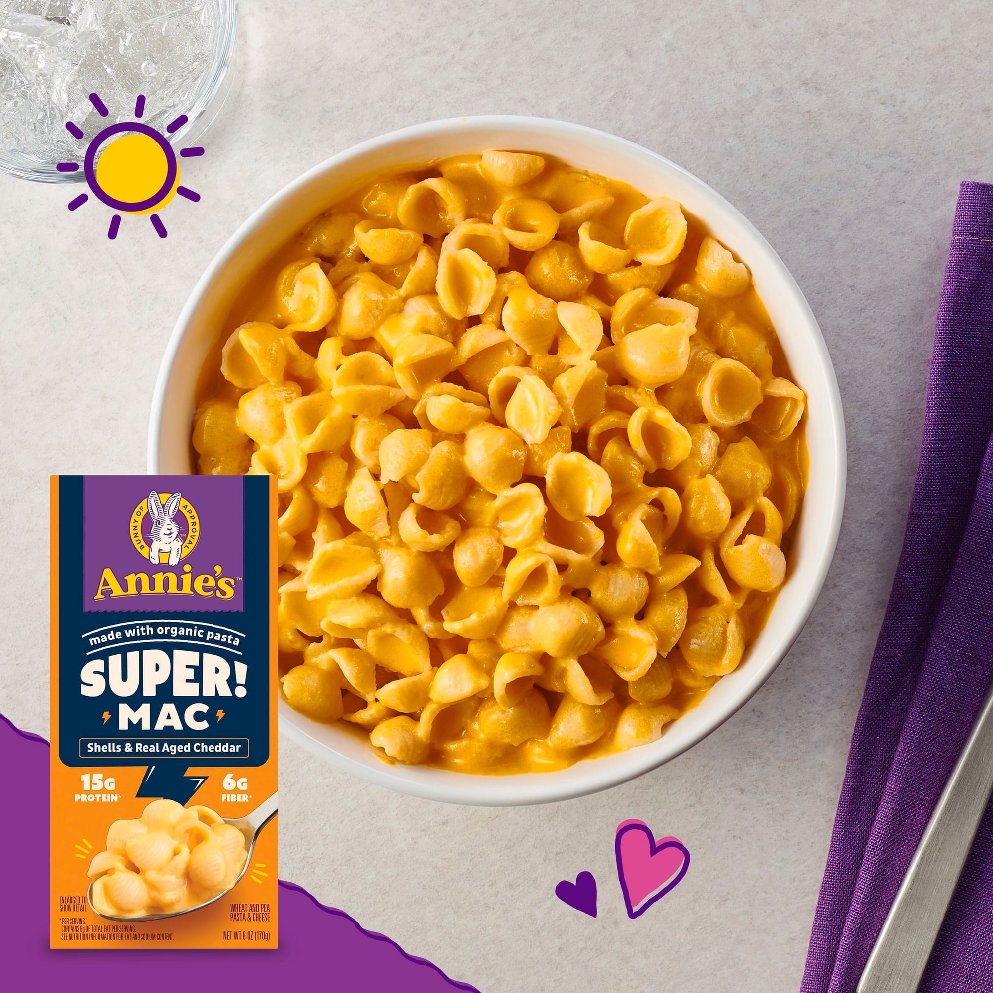 A bowl of creamy, orange shell pasta sits on a light surface next to a purple napkin and a glass of water, with a colorful box of Annie's Super Mac nearby, highlighting its organic ingredients.
