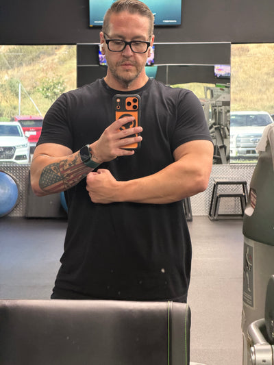 A man with glasses and a tattoo poses in a gym, holding a phone to take a selfie while standing in front of a mirror, showcasing his muscular arms and a focused expression.