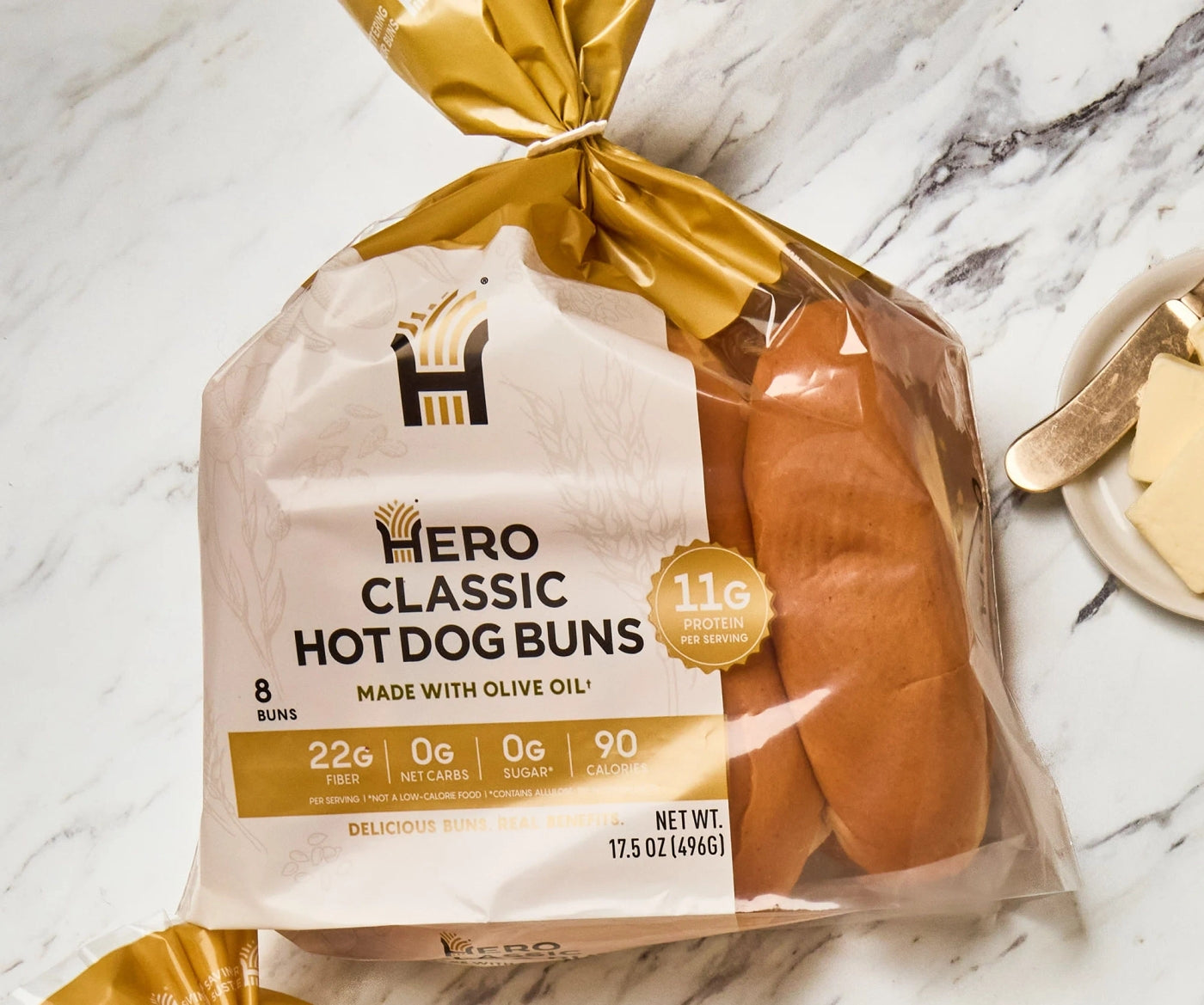A package of Hero Classic Hot Dog Buns, featuring eight buns made with olive oil, displays nutritional information and is set on a marble surface next to a plate with butter and a knife.