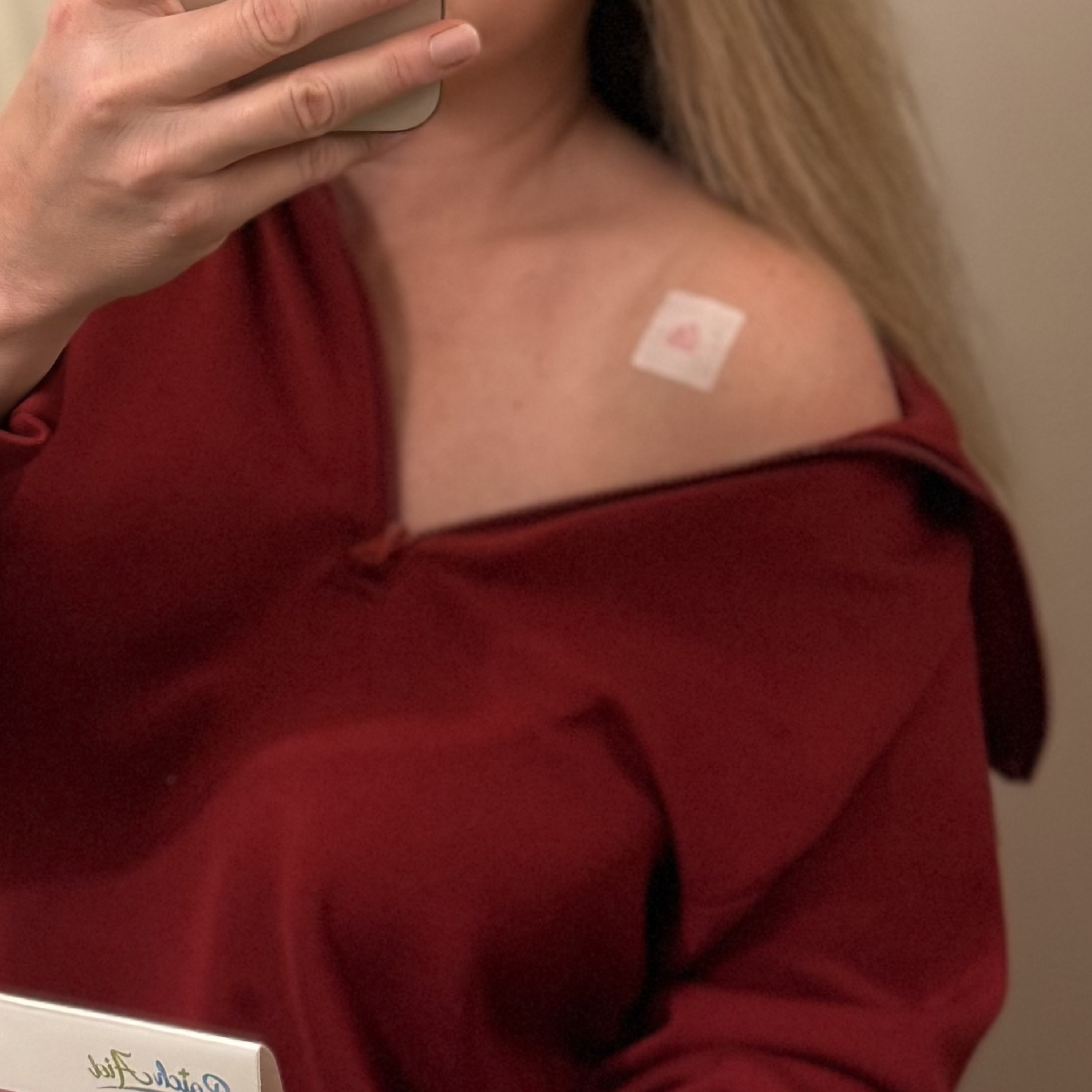 A person with long hair wearing a burgundy top takes a selfie, showing a small bandage on their shoulder and holding a product box in their hand.