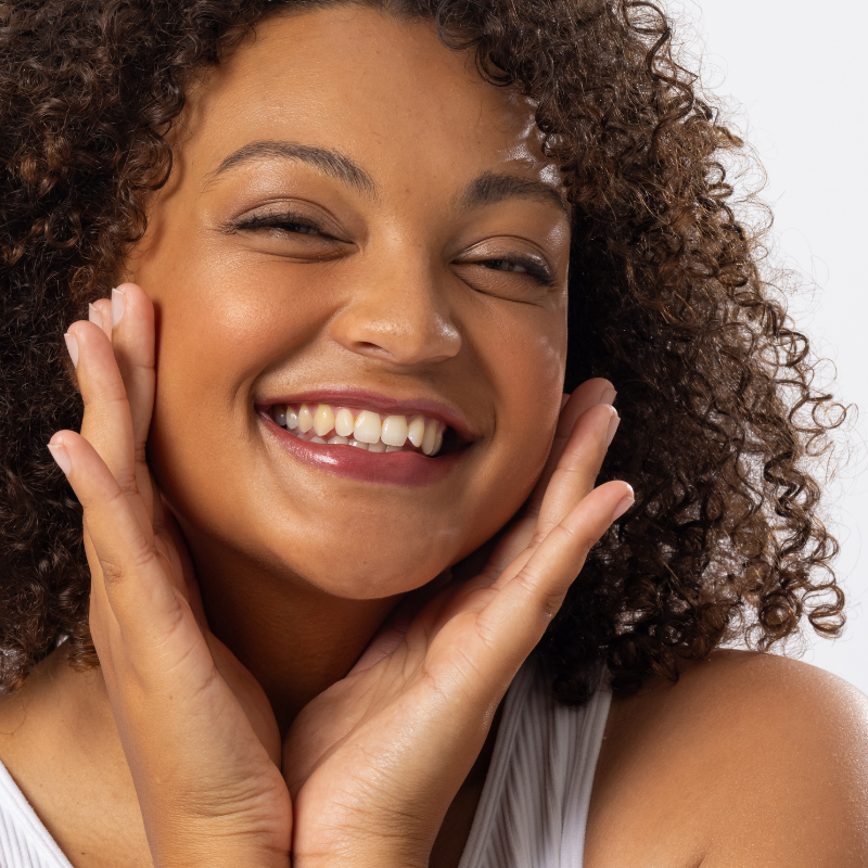 A smiling young woman with curly hair rests her hands on her cheeks, conveying joy and enthusiasm against a plain white background.