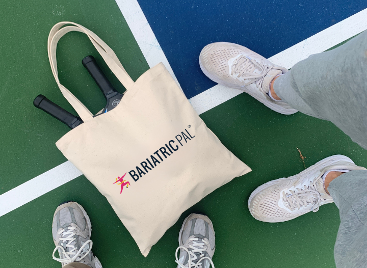 A beige tote bag with the logo "BARIATRICPAL" sits on a green and blue tennis court, with two pairs of athletic shoes visible nearby and tennis rackets peeking out of the bag.