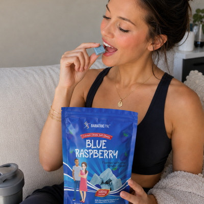 A woman in a black tank top is smiling while holding a blue raspberry calcium soft chew near her mouth, sitting on a couch with a bag of the product beside her.