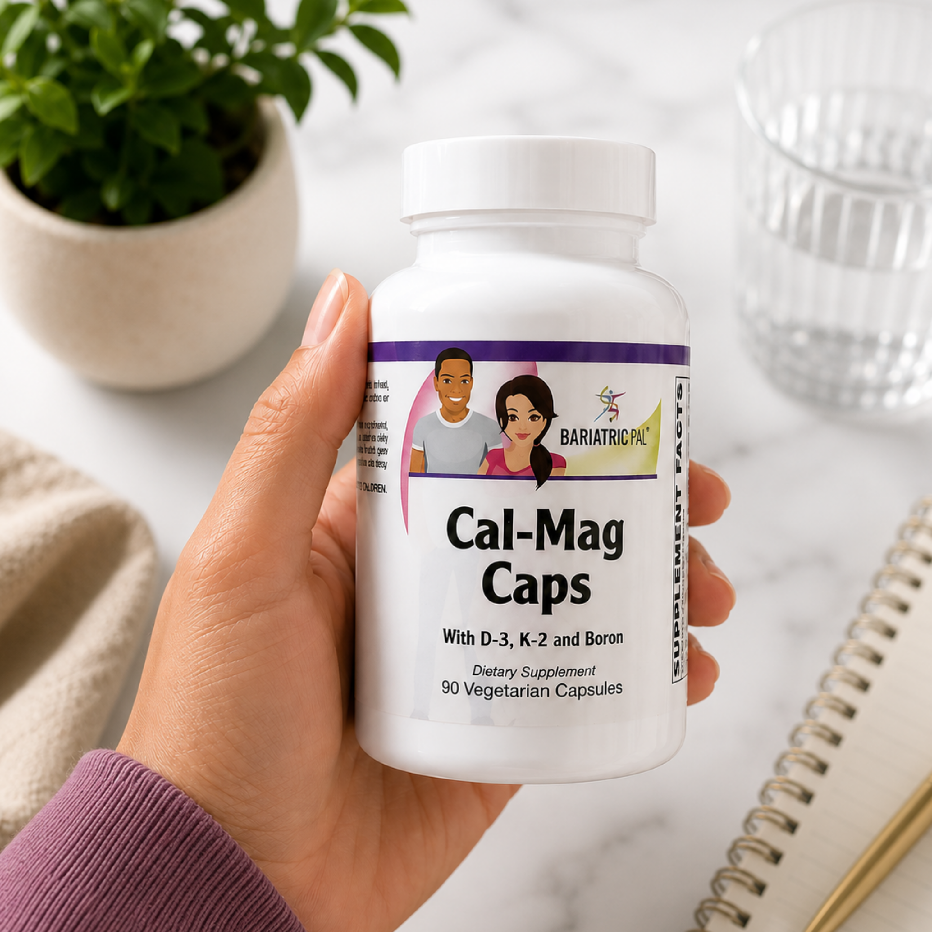 A hand holds a white bottle labeled "Cal-Mag Caps" with illustrations of two people, surrounded by a plant, a glass, and a notebook on a marble surface, suggesting a wellness or dietary context.