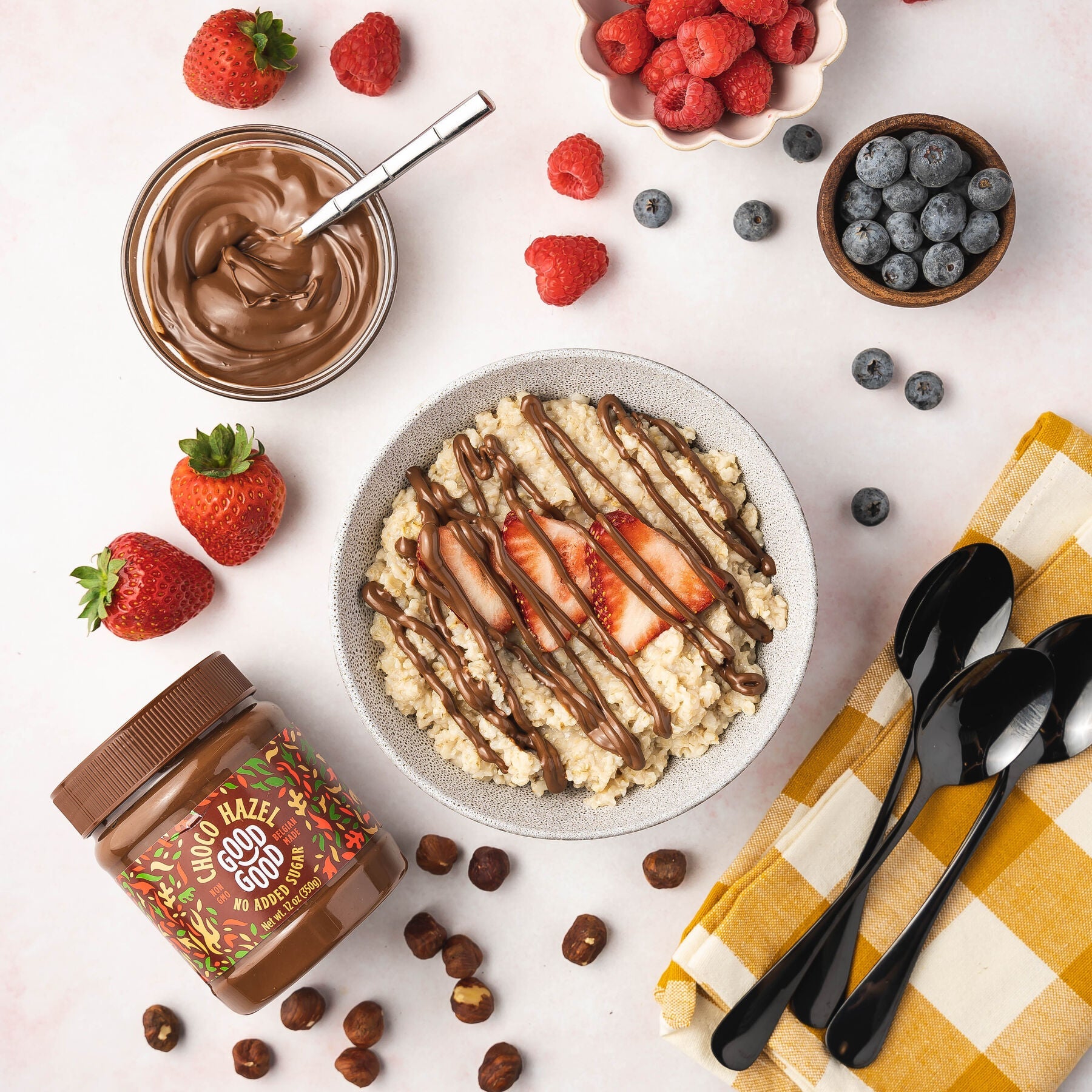 A bowl of oatmeal topped with sliced strawberries and drizzled with chocolate hazelnut spread is surrounded by fresh berries, a jar of hazelnut spread, and black spoons on a checkered napkin.