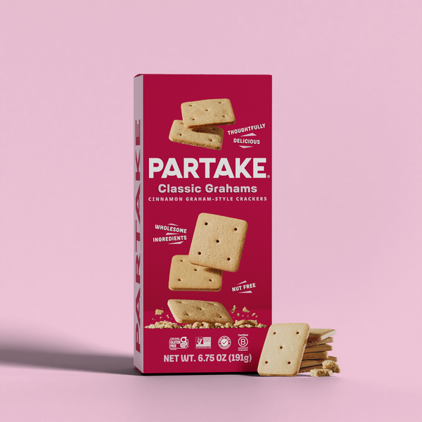 A box of Partake Classic Grahams, featuring cinnamon graham-style crackers, is displayed against a pink background with some crackers scattered beside the box.