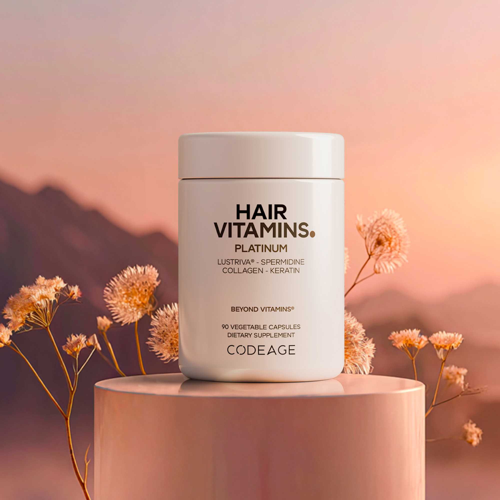 A white container labeled "HAIR VITAMINS. PLATINUM" with details about its ingredients stands on a round pedestal, surrounded by soft pink flowers and a pastel sunset backdrop.