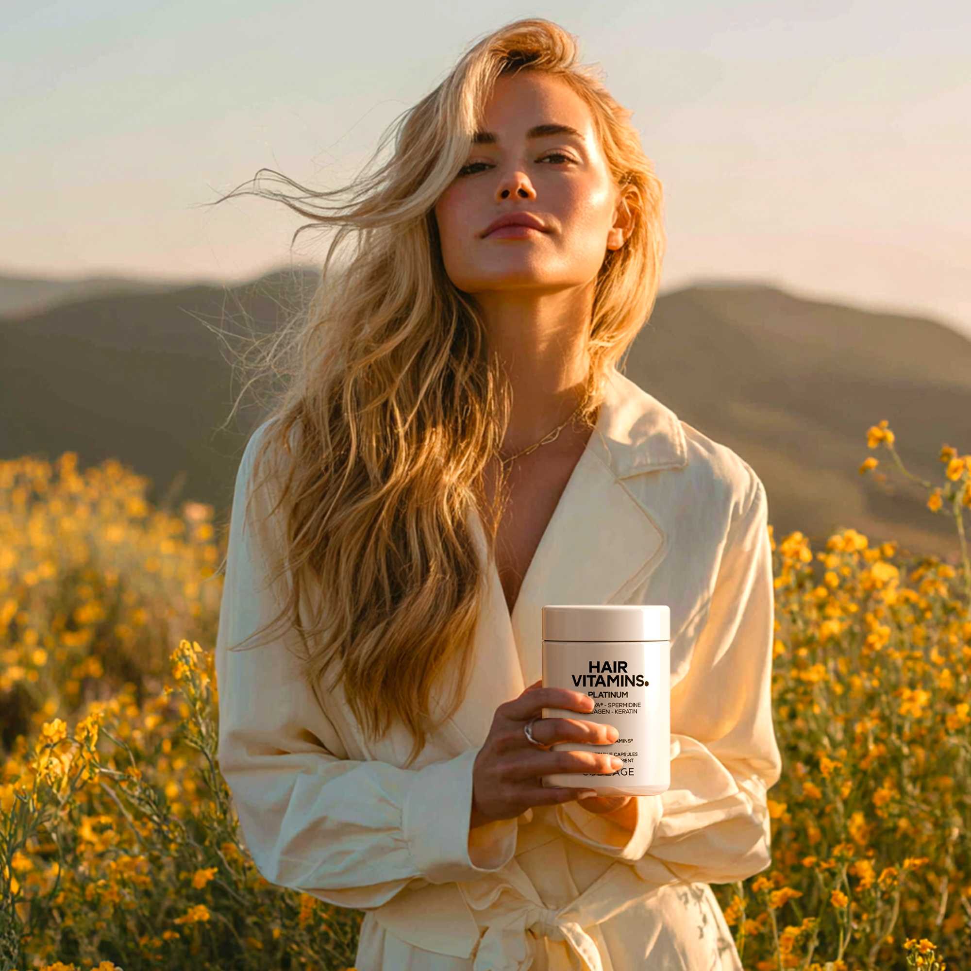 A woman with long, wavy blonde hair stands in a field of yellow flowers, holding a container labeled "HAIR VITAMINS," with mountains and a sunset in the background.