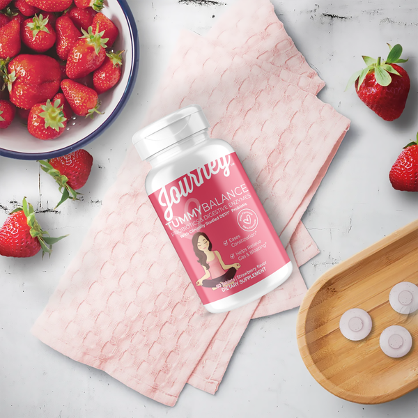 A bottle of Journey Tummy Balance probiotic supplement is placed on a pink cloth next to a bowl of fresh strawberries and a wooden tray with round, pink tablets.