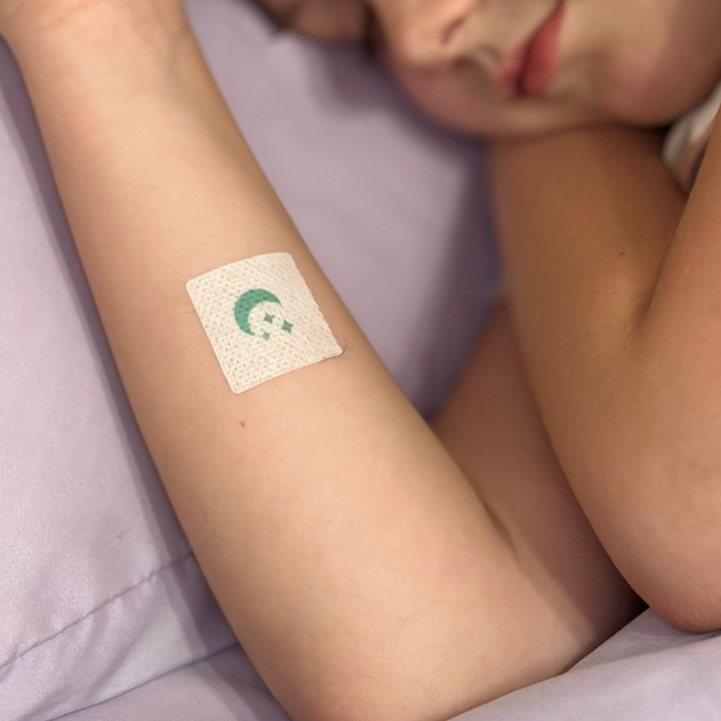 A child is resting with their eyes closed, and a colorful bandage is applied to their arm, resting on a soft, purple surface.