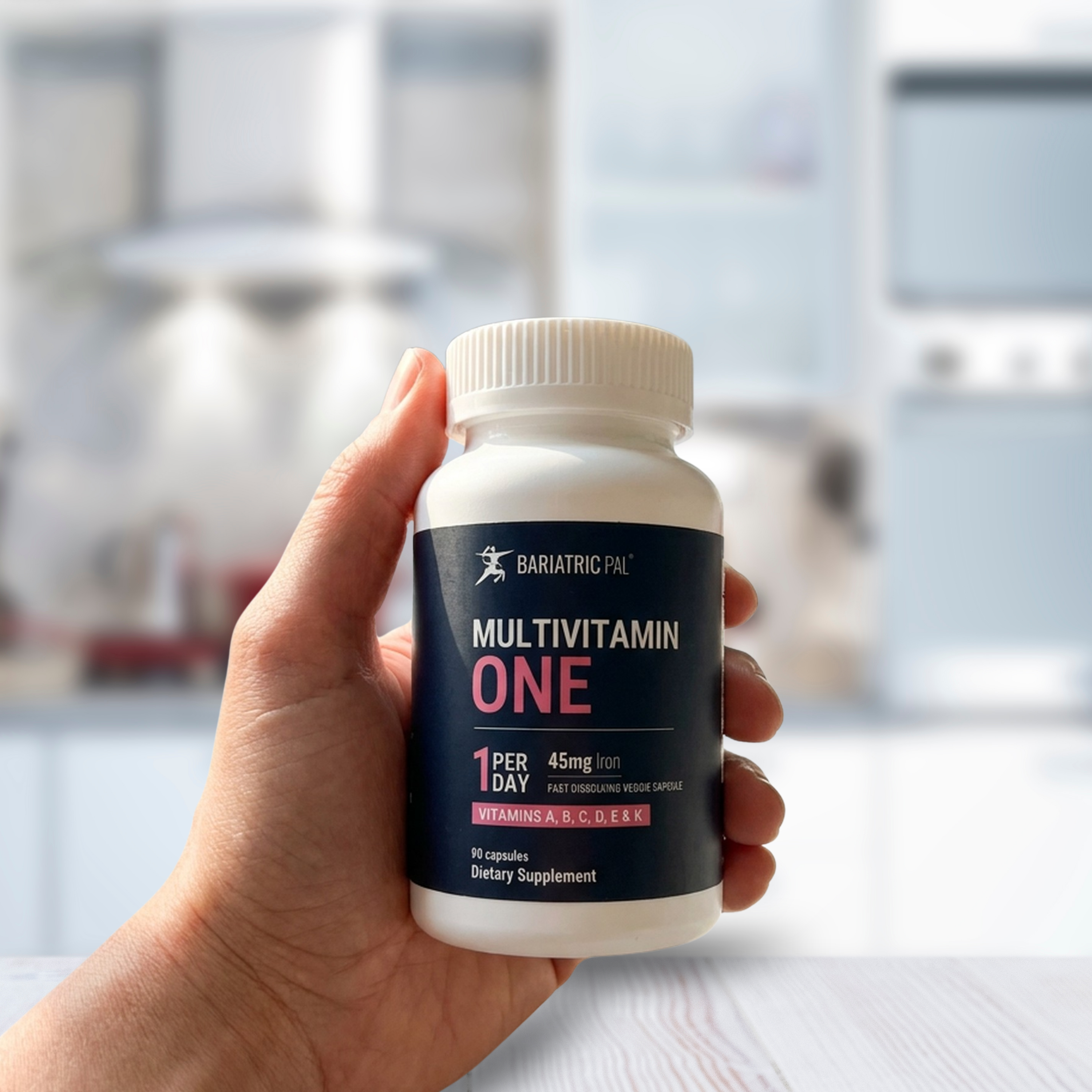 A hand holds a white bottle labeled "Multivitamin One" with details about daily dosage and vitamins, set against a blurred kitchen background.