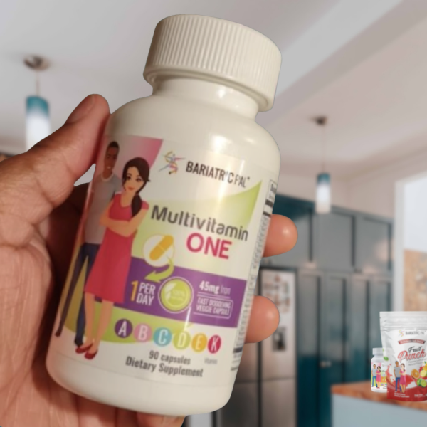 A hand holds a bottle of Bariatric Fusion Multivitamin One, featuring illustrations of a man and woman, in a modern kitchen setting with cabinets and other products visible in the background.