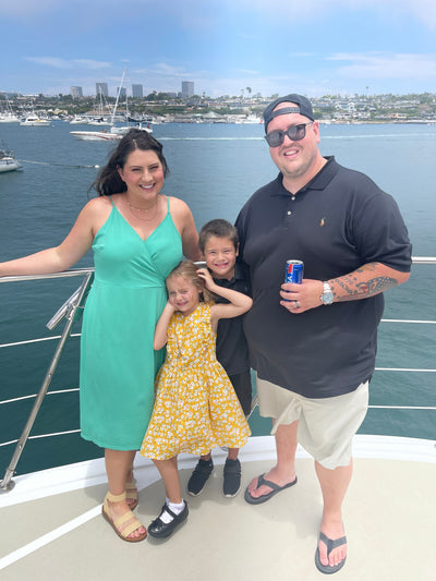 A woman in a green dress and a man in a black shirt stand on a boat with two children, smiling and enjoying a sunny day by the water, with boats and buildings visible in the background.