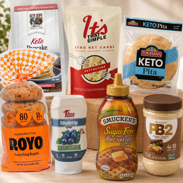A group of keto-friendly breakfast foods arranged together on a wooden surface, including ROYO Everything Bagels, That's That Simple Keto Pancake mix, TofuBay Keto Pita, Taste Blueberry Yogurt, Smucker's Sugar Free Breakfast Syrup, and a jar of PB2 Peanut Butter.