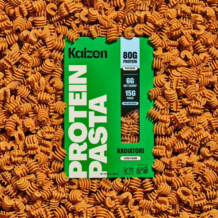 A box of Kaizen protein pasta is centered among a pile of radiatori pasta, featuring bold green packaging with nutritional information highlighting 80g of protein, 6g net carbs, and 15g fiber per serving.