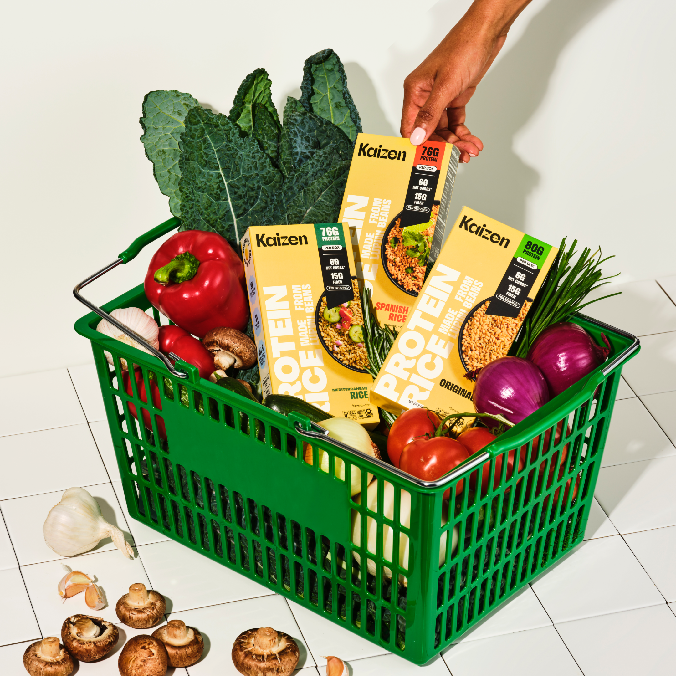 A green shopping basket filled with colorful vegetables, including peppers and mushrooms, alongside three boxes of Kaizen protein rice, sits on a tiled surface.