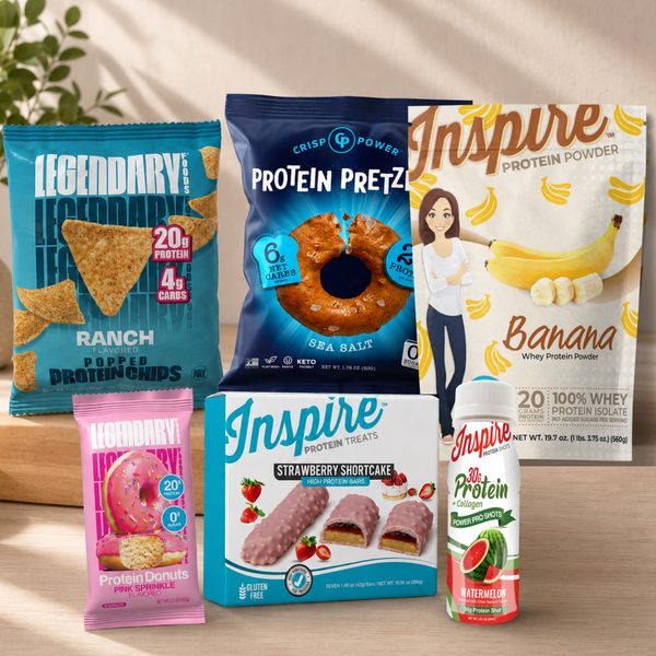 A grouped assortment of snack and protein products arranged on a wooden surface against a warm beige background. Visible items include a bag of Legendary Ranch Popper Protein Chips, a blue bag of Crisp Power Protein Pretz Sea Salt, a large white bag of Inspire Protein Powder Banana flavor, a pink bag of Legendary Protein Cookies Pink Glazed, a blue box of Inspire Protein Treats Strawberry Shortcake, and a white and red bottle of Inspire 30 Protein Watermelon flavor.