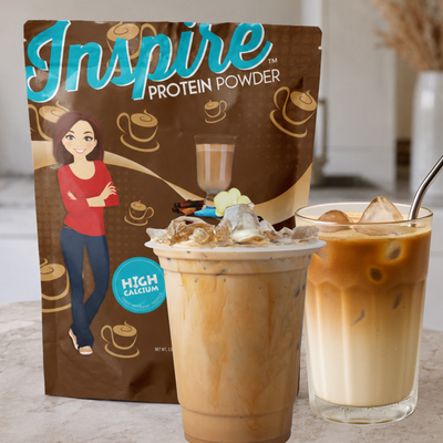 A bag of Inspire protein powder featuring a woman with crossed arms is displayed alongside two iced coffee drinks on a countertop, highlighting the product's high calcium content.