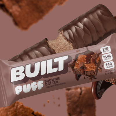 A close-up of a chocolate-coated protein bar labeled "Built Puff Brownie Batter," with a bite taken out, surrounded by brownie pieces and a smooth chocolate drizzle on a brown background.