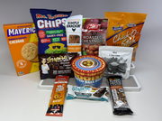A variety of packaged snacks and food items, including chips, protein bars, soup, and nuts, are arranged on a white surface, showcasing their colorful packaging and labels.