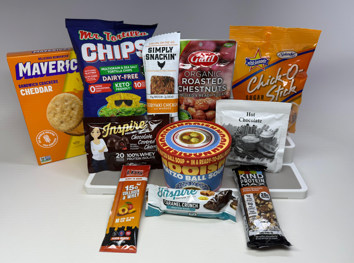 A variety of packaged snacks and food items, including chips, protein bars, soup, and nuts, are arranged on a white surface, showcasing their colorful packaging and labels.