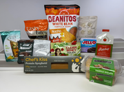 A variety of packaged food products are displayed on a shelf, including snacks, protein items, and sauces, highlighting options like nacho tortilla chips and gluten-free spaghetti.