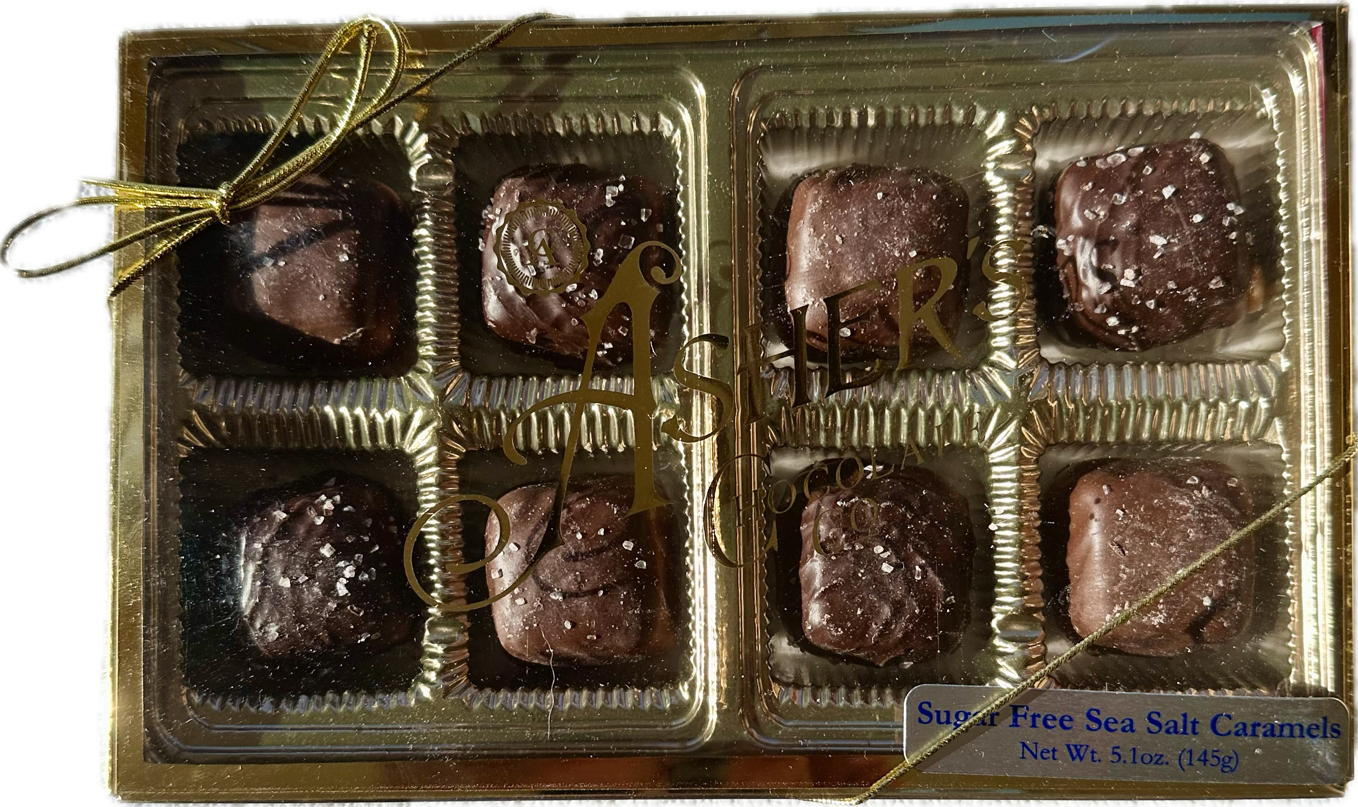 A gold box tied with a ribbon contains eight chocolate-covered caramels sprinkled with sea salt, labeled as "Sugar Free Sea Salt Caramels" from Asher's Chocolate Co.