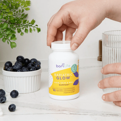 A hand reaches for a white bottle labeled "Bariatric Glow" on a marble countertop, accompanied by a bowl of blueberries and a glass of water, suggesting a healthy lifestyle.