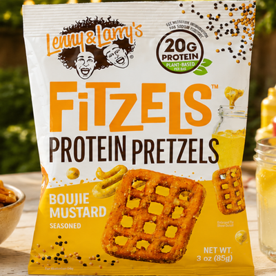 A bag of Lenny & Larry's FitZels Protein Pretzels in boujie mustard flavor is displayed on a wooden table, featuring vibrant branding and a background of pretzels and mustard.