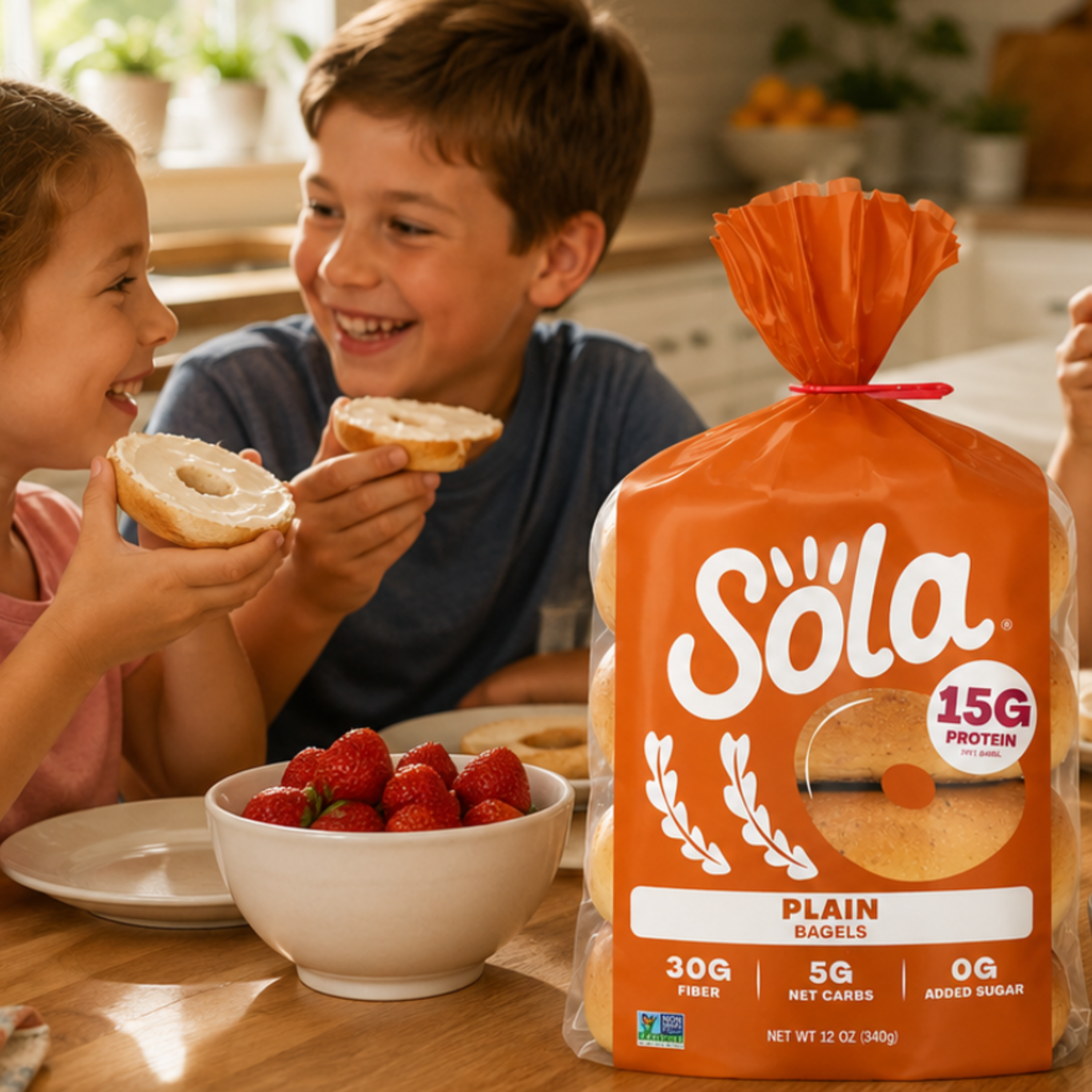 A large bag of Søla Plain Bagels sits on a kitchen table next to a bowl of strawberries, with two children holding bagel halves in the background.