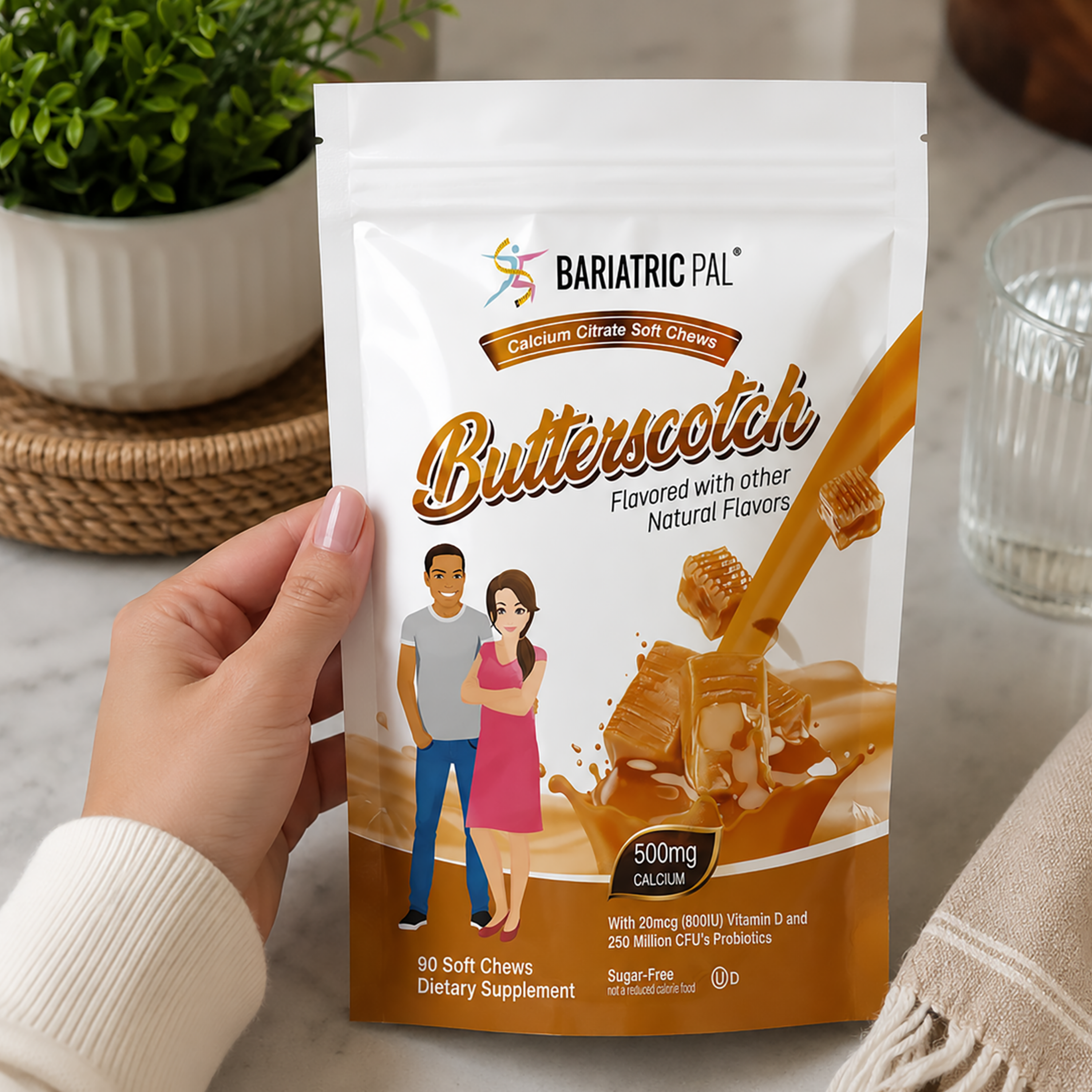 A hand holds a stand-up pouch of BariatricPal Butterscotch soft chew dietary supplement. The white and brown package features illustrations of a man and woman and a butterscotch splash graphic.
