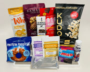 A variety of protein-rich snacks is displayed, including cheddar cheese crisps, protein popcorn, pretzels, oatmeal, and Jell-O, arranged on a white surface against a plain background.