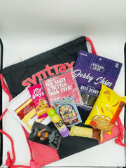 A black drawstring bag with red accents contains various snacks, including beef jerky, protein bars, and chocolate treats, arranged attractively against a light background.