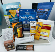 A variety of protein-rich snacks and beverages, including jerky chips, protein chips, coffee, and macaroons, are arranged on a white tray against a light background.